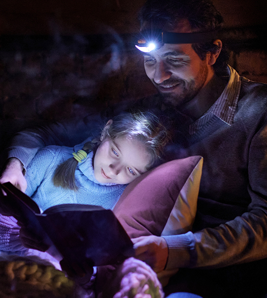 Dad reading to his daughter in the dark with a headlamp during a power outage.