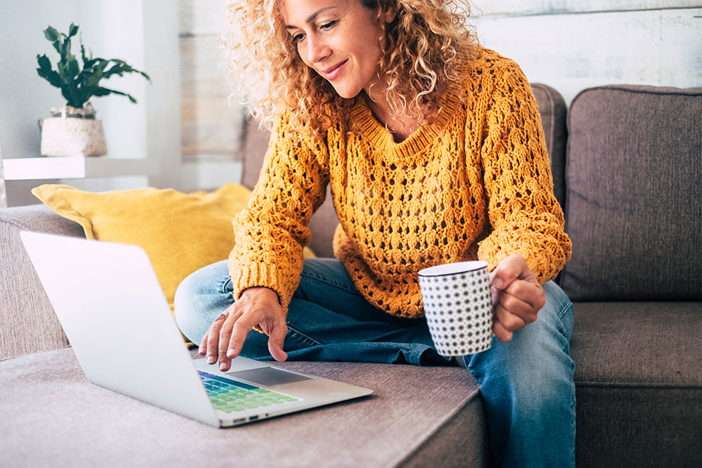 Woman wearing yellow sweater using laptop on couch while drinking coffee