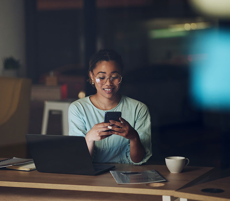 Woman seated in front of laptop in the dark, looking down at mobile phone