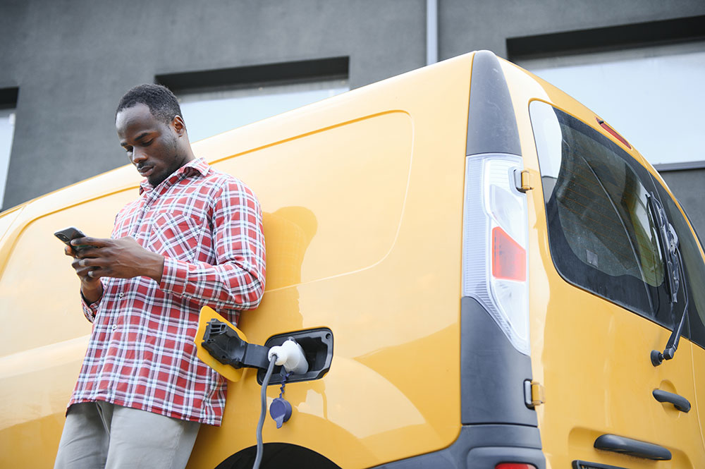 African American man reviewing his phone beside a yellow EV delivery van as it charges