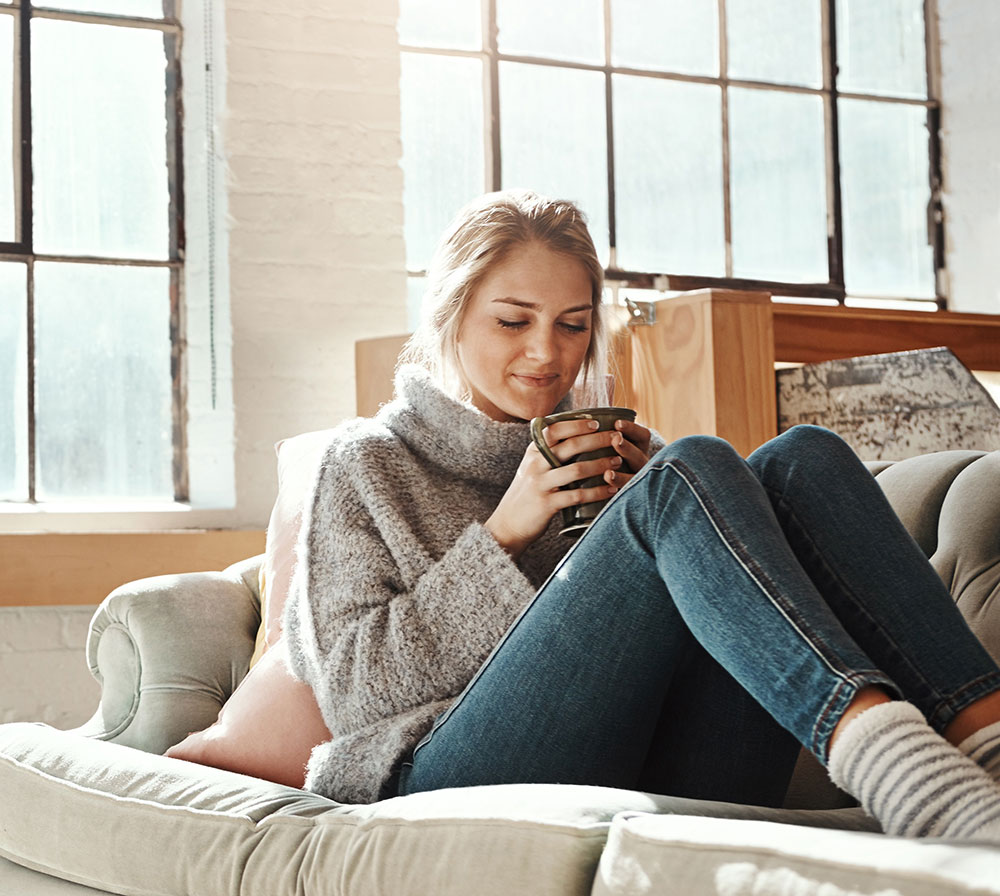 Woman cuddled up on couch at home with bright sun shining through windows
