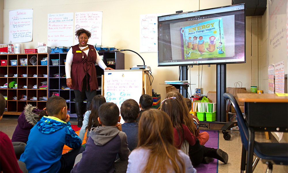 De’Kendrea reads to a group of students in a classroom with their backs turned to the camera.