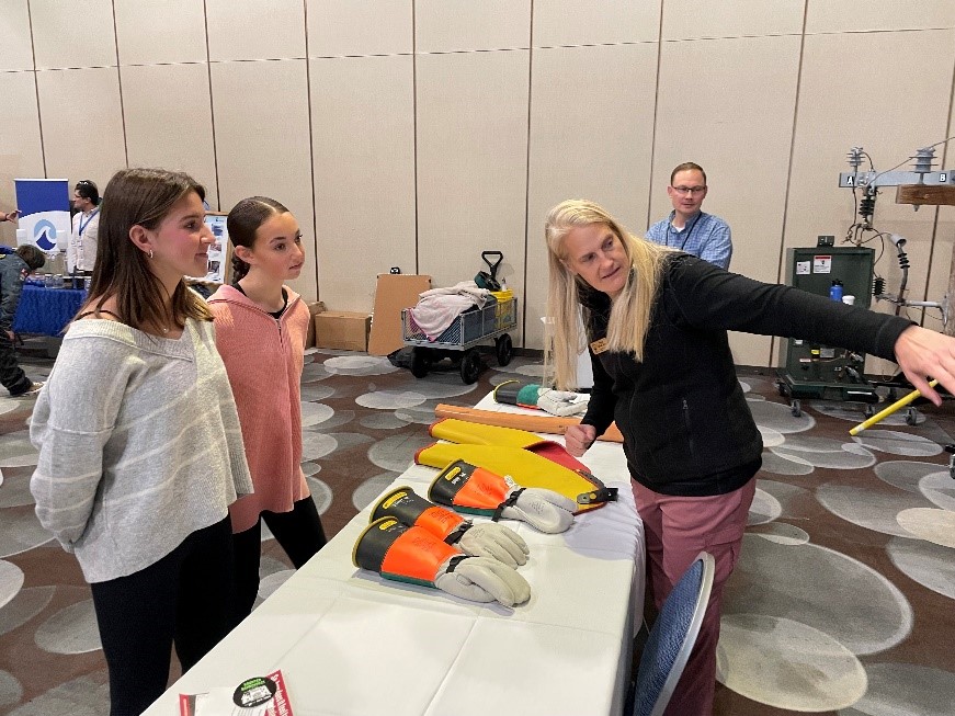 A woman talks to two female students as she points. There's a table with gloves in between them.