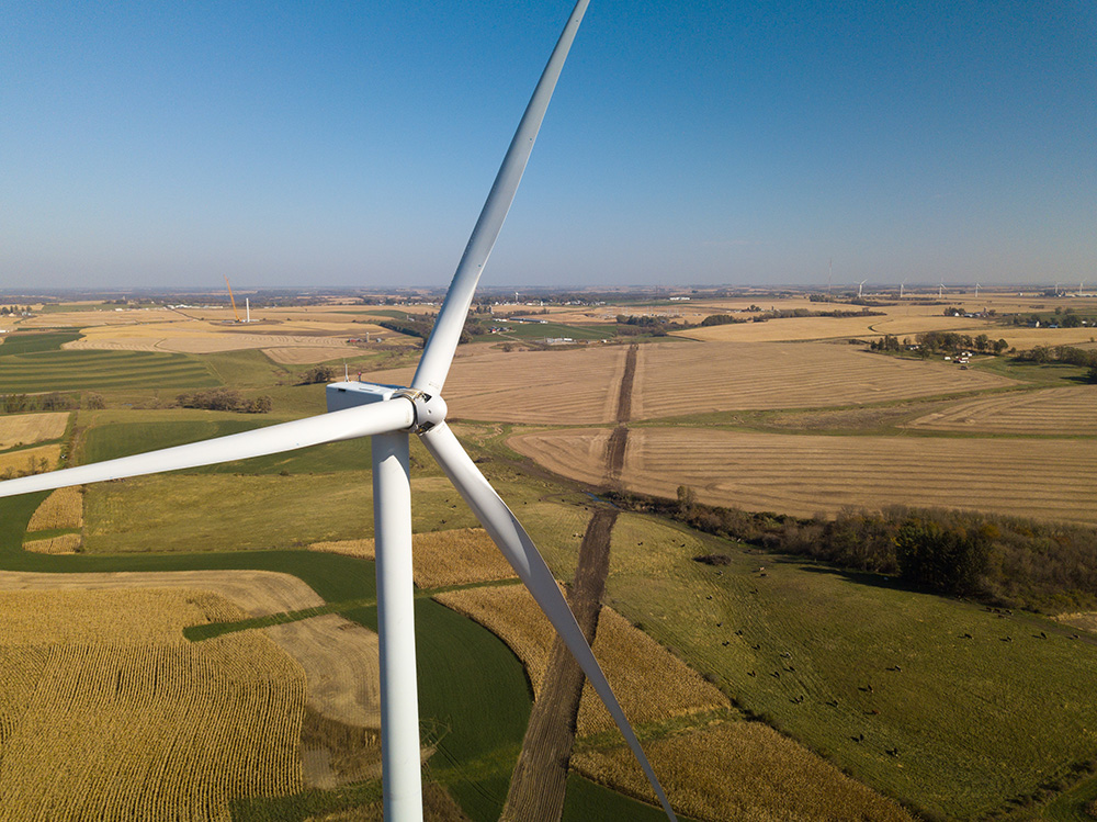 Aerial view of a wind turbine. Blue sky above and green and brown fields below.