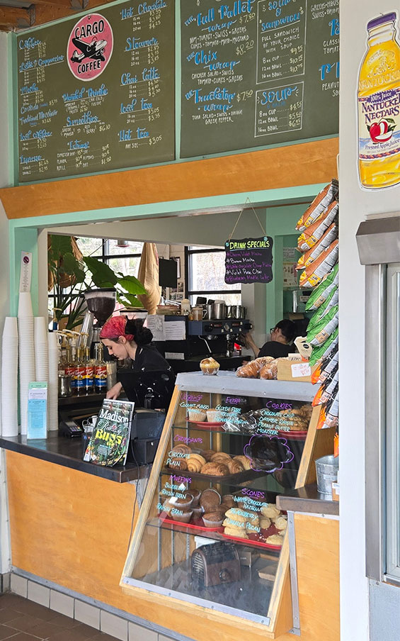 The counter at Cargo Coffee, with treats in the case, stacked cups and a chalkboard menu above.