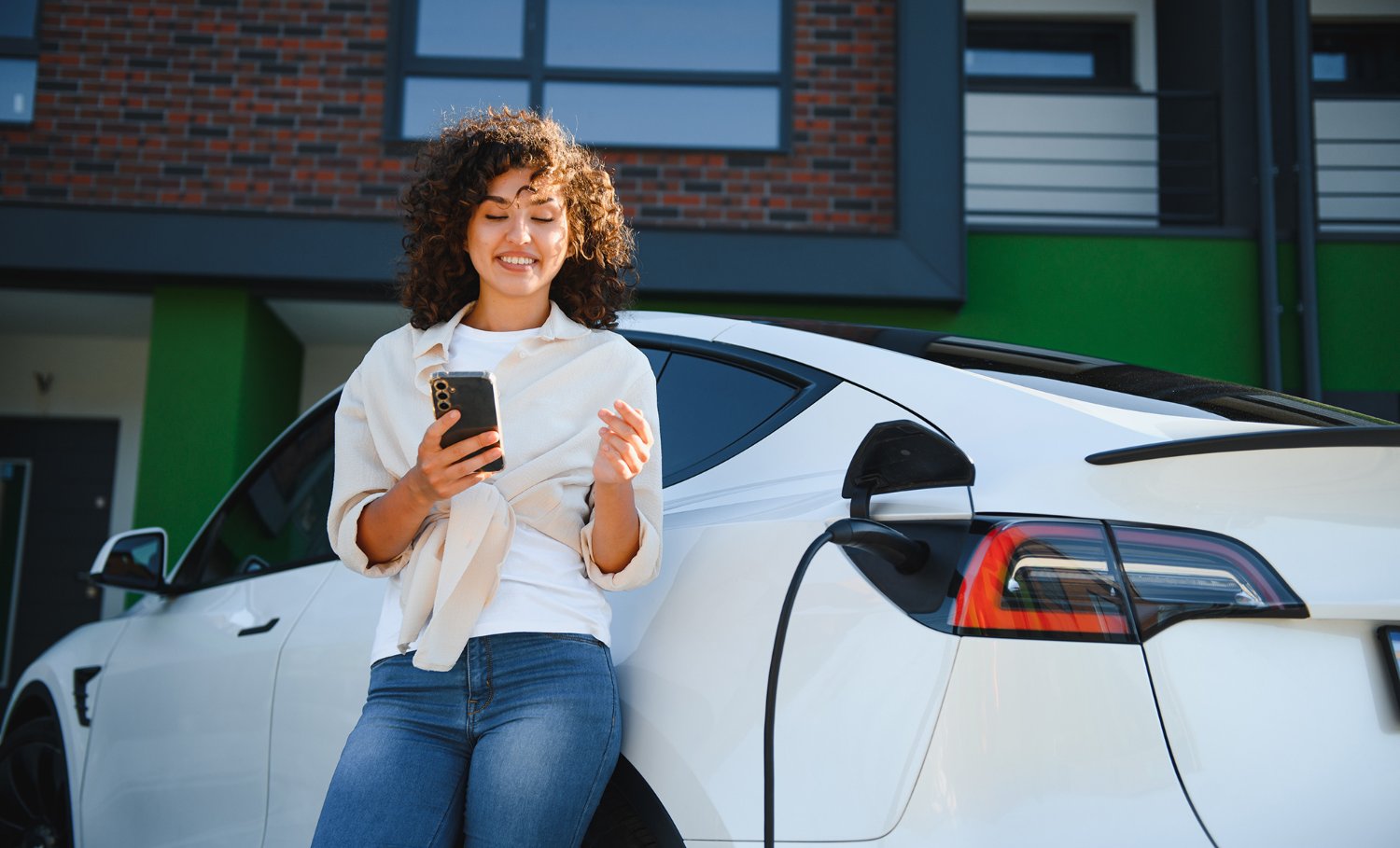 Woman leaning on car, looking at phone while charging electric vehicle