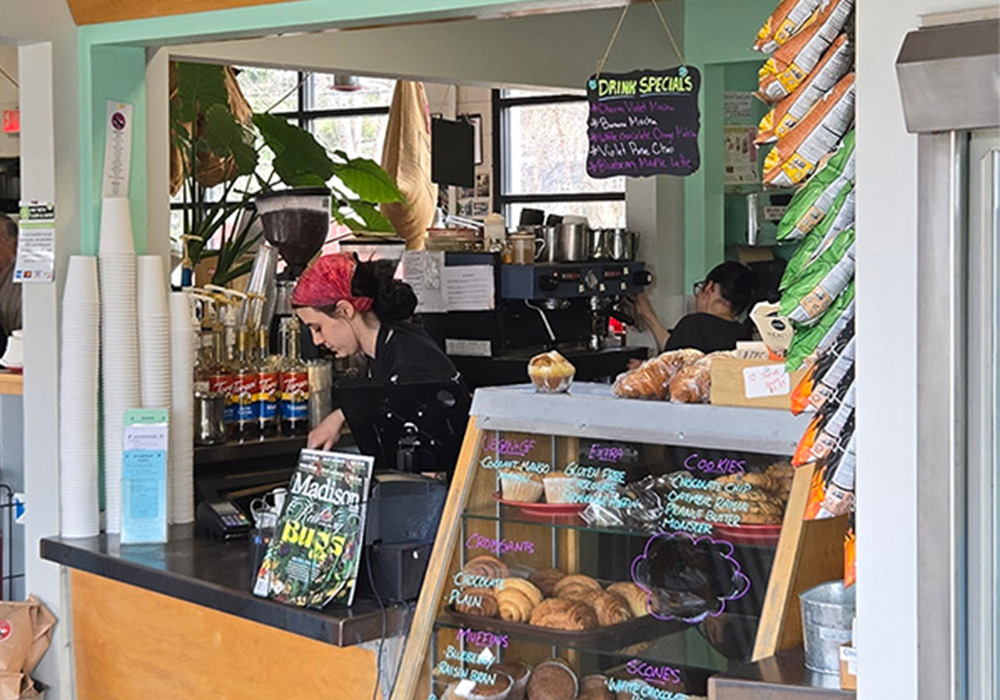 The front counter at Cargo Coffee, with two women working, a stack of white coffee cups and treats behind the window display.