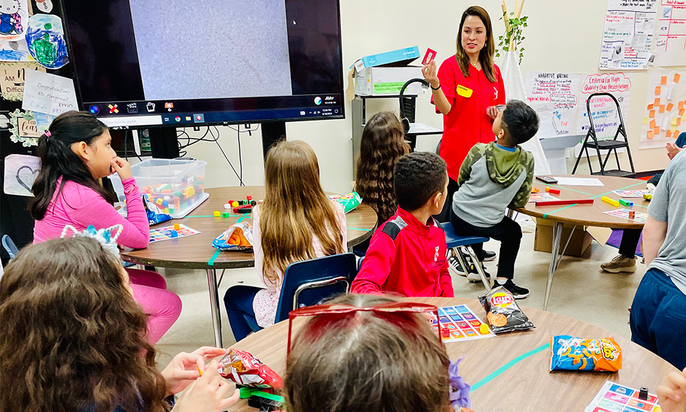 Gloria holds up a card to play La Lotería de la Energía with students seated at circular tables.
