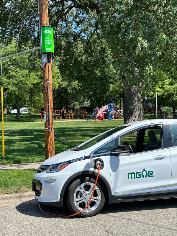 A white electric vehicle parked curbside, plugged into a pole-mounted EV charger.