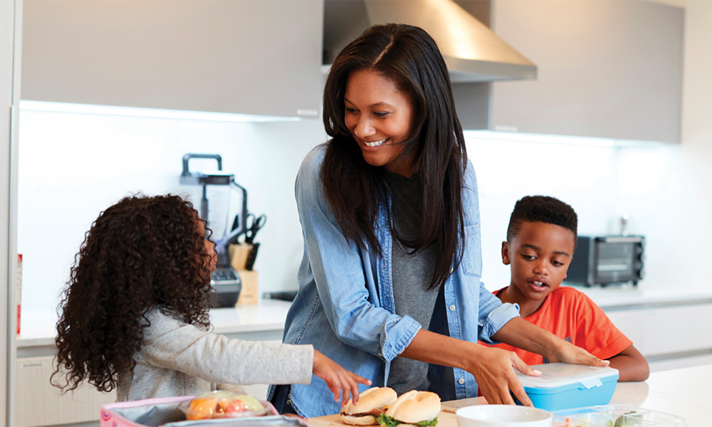 An African American mom, son and daughter standing by a kitchen island as she packs their lunches.
