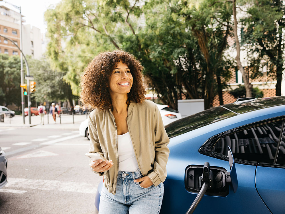 Smiling woman leaning on her car while it charges at an EV charging station
