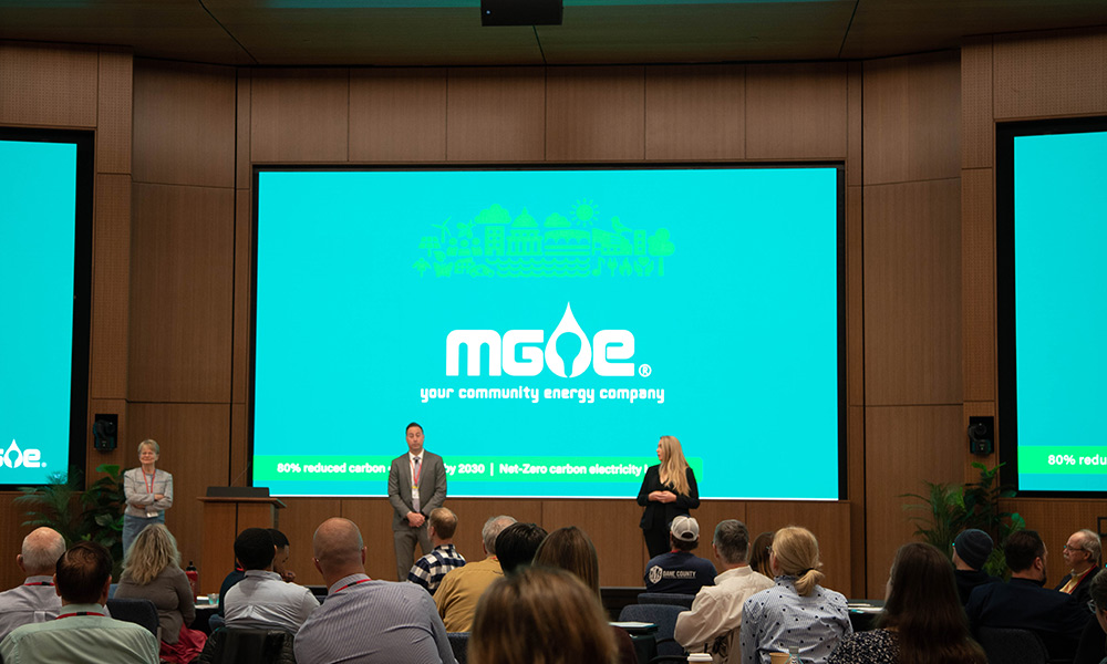 A back-of-the-room view of the 2024 Business Customer Forum, with the backs of people at tables, three large screens with a turquoise background and two presenters.
